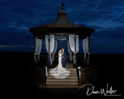 A newlywed couple sharing a romantic moment under a beautifully lit gazebo against a dusky sky.