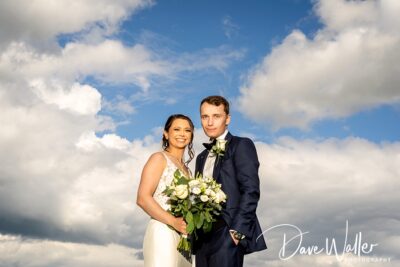 A couple dressed in wedding attire, sharing a moment under a sky painted with fluffy clouds.