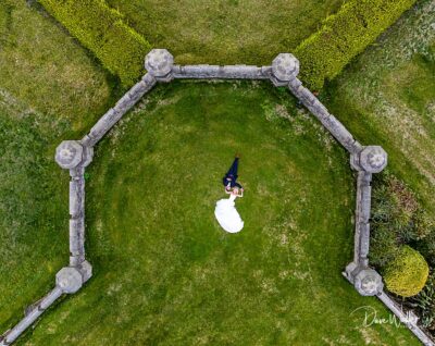 Aerial view of a bride in a white dress standing on a lush green lawn in West Yorkshire, surrounded by an ornate stone hedge, creating a striking contrast and a moment of serenity by the