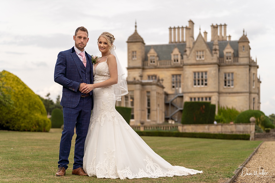 A bride and groom holding hands, sharing a joyful moment, with the elegant Stoke Rochford Hall in the background.