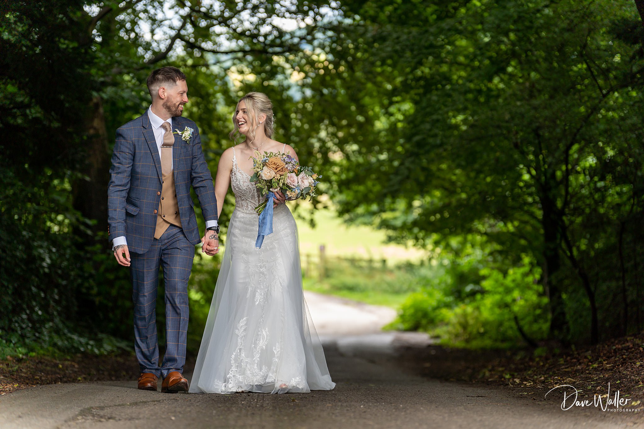 Danielle & David, a joyful bride and groom holding hands and walking down a sun-dappled country lane at East Riddlesden Hall, surrounded by lush greenery, sharing a moment of laughter