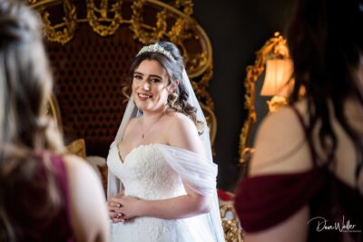 A radiant bride shares a joyful moment with someone off-camera, her happiness reflected in a mirror within an elegantly furnished room.
