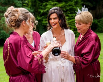 Three women in elegant robes and dressed hair sharing a joyful moment with a toast outdoors.