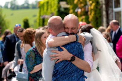 Two men sharing an emotional embrace at a sunny outdoor gathering, surrounded by guests in celebration attire, conveying a poignant moment of connection and joy.