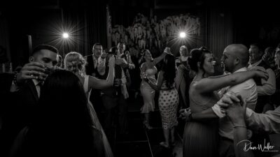 An elegant black and white snapshot capturing a joyful moment with couples dancing and celebrating at a festive event, illuminated by dramatic backlighting.