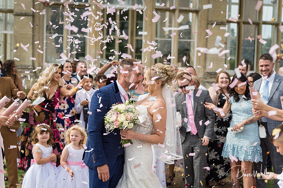 A joyful bride and groom share a kiss amidst a shower of confetti with smiling guests celebrating around them at their wedding at Stoke Rochford Hall.