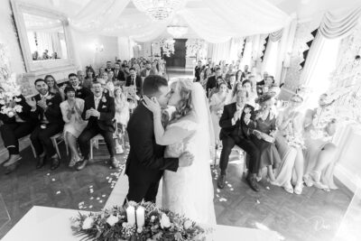 A bride and groom share a romantic kiss at the altar as their wedding guests joyfully look on, capturing a timeless moment of love and celebration.