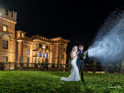 A joyful newlywed couple celebrating their wedding night with a champagne spray in front of a majestic castle illuminated against the night sky.