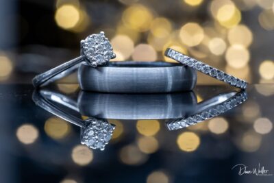 A pair of wedding rings elegantly displayed, with the engagement ring featuring a sparkling diamond cluster, and the wedding band showcasing a subtle texture, both reflecting on a glossy surface against a backdrop of golden bokeh lights.