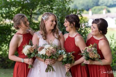 A joyful bride in a white dress shares a heartfelt laugh with her bridesmaids, who are dressed in elegant red gowns, while they all hold beautiful bouquets in a lush outdoor setting.
