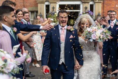A jubilant newlywed couple gleefully walks through a shower of confetti, surrounded by the laughter and cheers of friends and family on their wedding day.