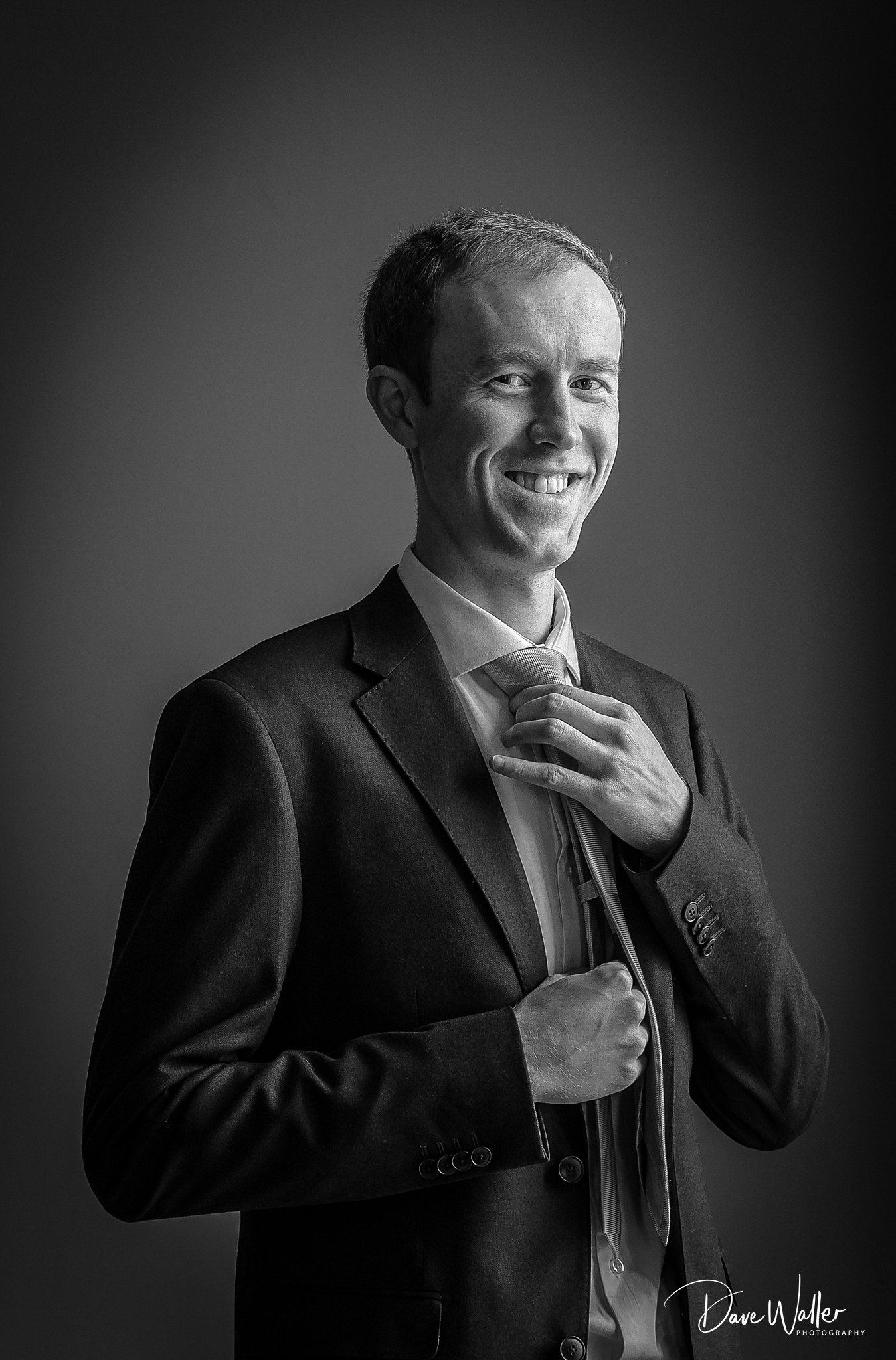 A monochrome portrait of a confident man in a suit, smiling and casually adjusting his tie at Rudding Park.