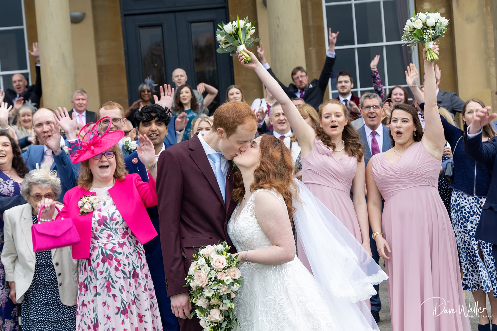 A joyful newlywed couple, Natasha & Marcus, share a kiss as they are surrounded by excited friends and family cheering and celebrating the special moment at their Rudding Park Wedding.