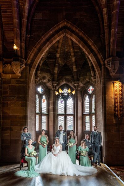 Elegant wedding party posed in a grand, gothic hall in West Yorkshire with stained glass windows, the bride seated at the center, exuding timeless grace and charm.