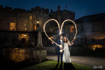 A couple celebrates their love with sparklers, creating a heart-shaped light trail, against the backdrop of an enchanting castle at dusk as they maintain eye contact.