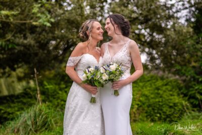 Two brides sharing a joyous moment, surrounded by nature, holding beautiful bouquets and gazing into each other's eyes with smiles of love and happiness.