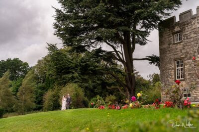 A couple in wedding attire holding hands in the distance, with vibrant flowers in the foreground and an imposing tree and stone building as a backdrop, creating a serene and romantic atmosphere.