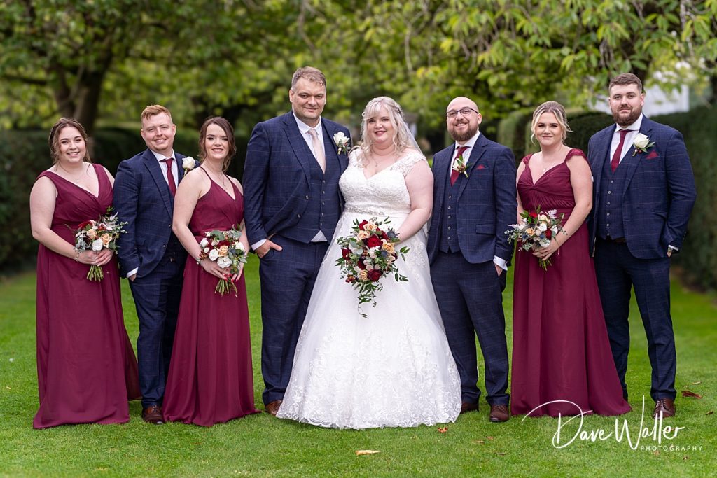 A joyful bridal party posing for a photo in Hazlewood Castle's garden, with the happy couple Kirsty & David in the center surrounded by bridesmaids in matching burgundy dresses and groomsmen