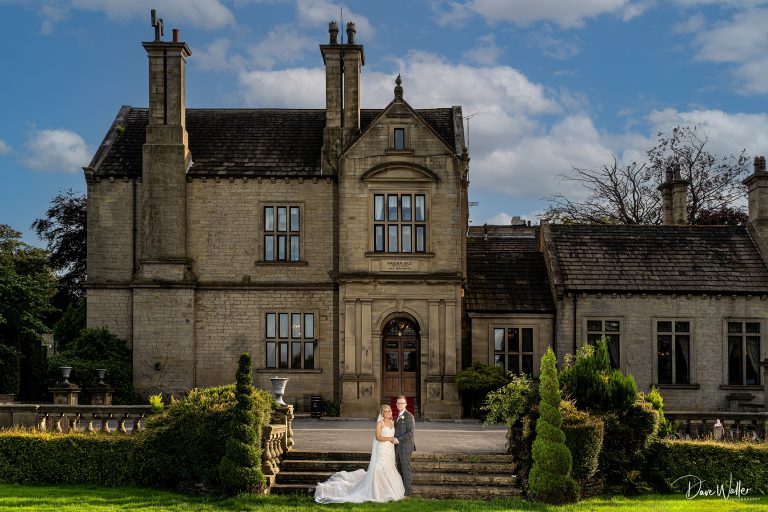 Lorna & Daniel, a newlywed couple, sharing a tender moment in front of the elegant Bagden Hall Hotel under a bright blue sky.