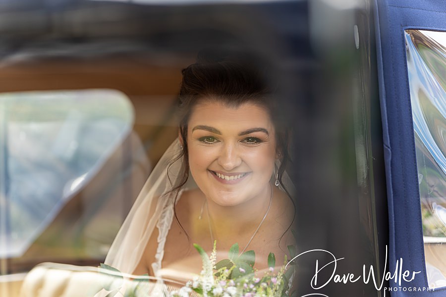 A radiant bride smiles warmly from the backseat of a car, exuding happiness and anticipation at her Sandburn Hall wedding.