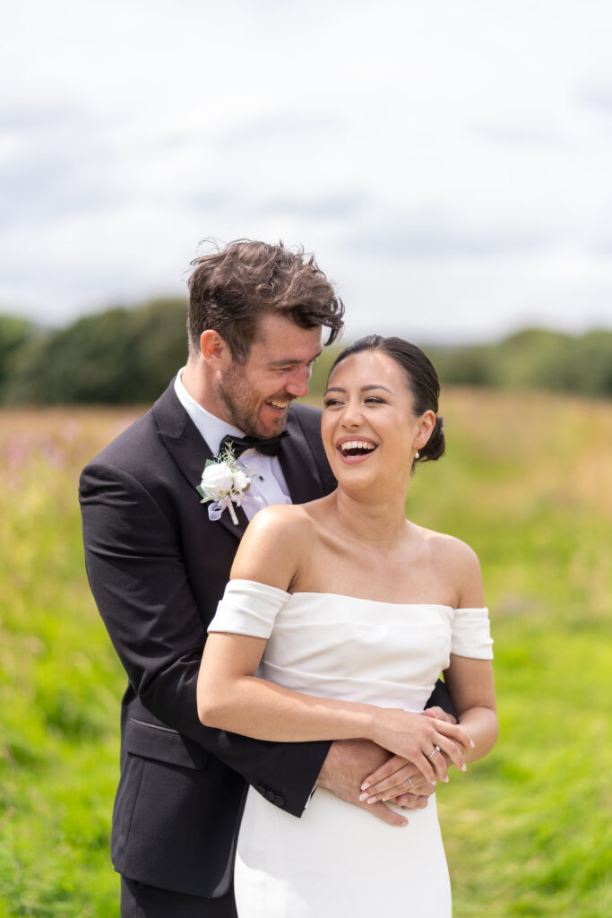 A joyful couple in wedding attire embraces and laughs in a grassy field, perfectly captured in this stunning piece of wedding photography. The groom wears a black suit with a boutonniere, and the bride dazzles in her white off-shoulder gown against blurred greenery and a cloudy sky, fit for any gallery.