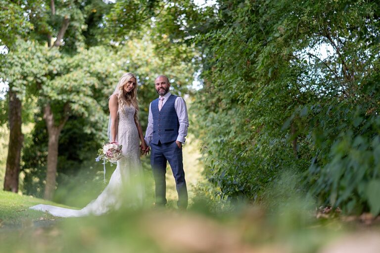 A couple stands outdoors amidst lush greenery during their wedding. The woman wears a long, lace gown and holds a bouquet, while the man sports a vest and tie. Sunlight filters through the trees, creating a serene and romantic atmosphere perfect for photography in this natural gallery.