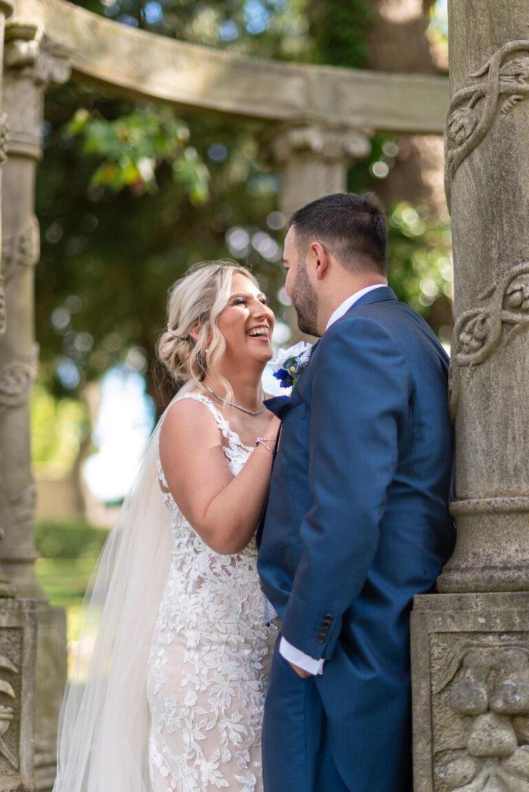 A smiling bride in a lace wedding dress leans against a groom in a blue suit under a stone gazebo adorned with intricate carvings. The lush greenery creates a serene atmosphere, perfectly captured for our wedding photography gallery.