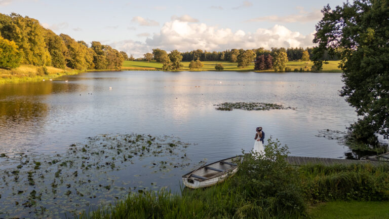 A serene lake surrounded by lush trees under a cloudy sky creates the perfect backdrop for wedding photography. A lone person in a white dress stands on a dock near a small boat, gazing at the water. Lily pads float near the shore, adding to this gallery-worthy peaceful scene.
