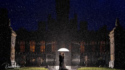 A couple shares a romantic moment under an umbrella as they stand before an illuminated, majestic castle at night, with raindrops surrounding them, creating a magical atmosphere, indifferent to the prices of admission into this