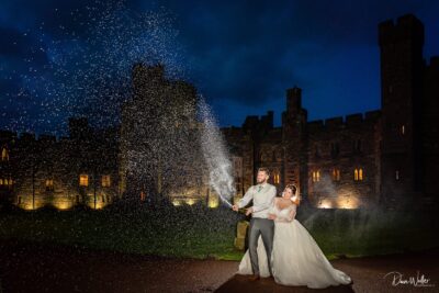 A newlywed couple sharing a magical moment during their recent wedding, creating a sparkling fountain of champagne under the night sky, with the grandeur of an ancient castle illuminated in the background.