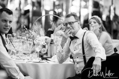 A black-and-white photo shows a man in a bow tie and suspenders sitting thoughtfully at a dining table set with wine glasses and plates. Another person beside him looks on, while a woman in the background appears to be speaking. The image, ideal for any Wedding Gallery, is signed "Dave Waller Photography.