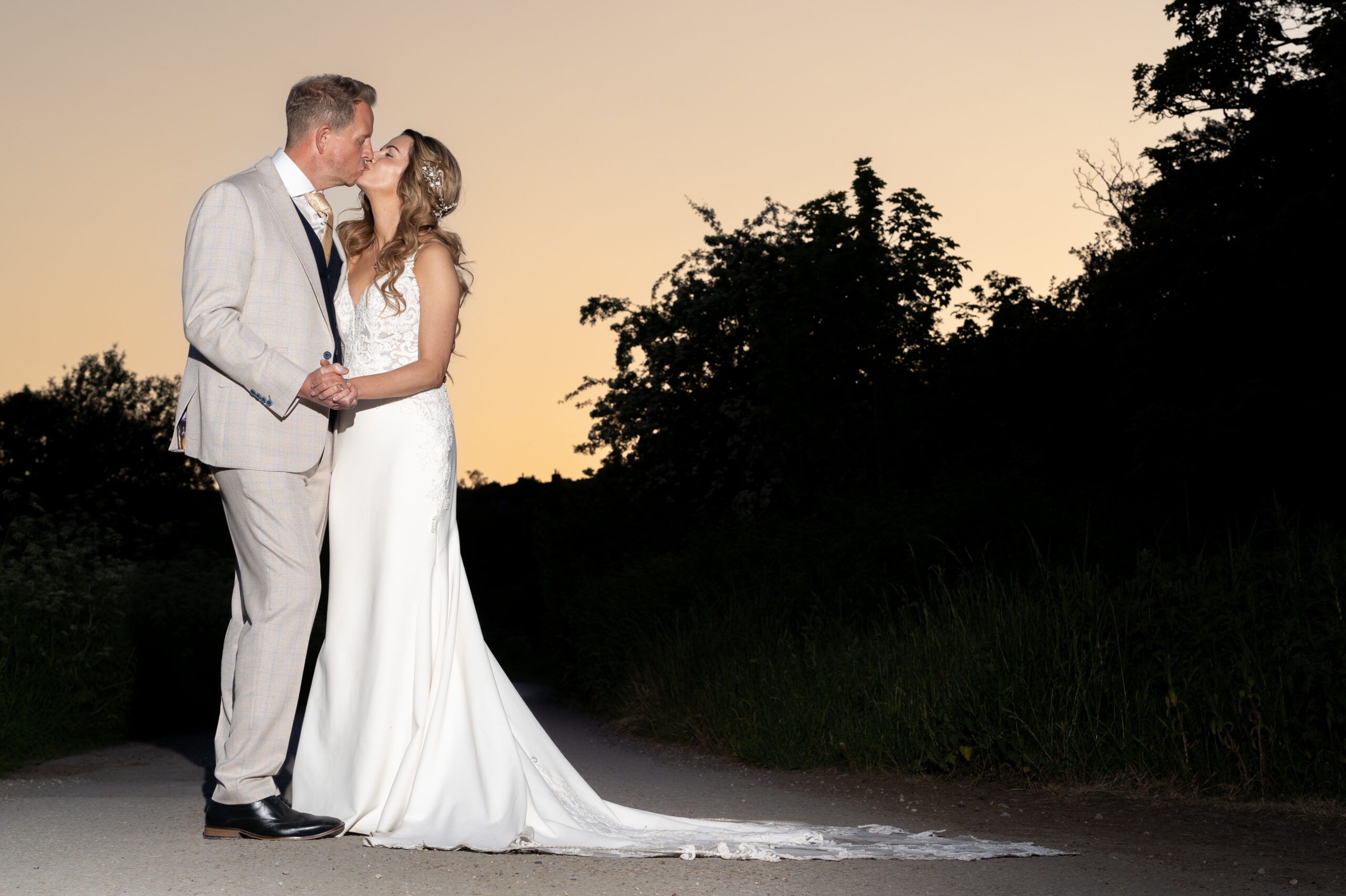 Bride and groom kissing at sunset on path.