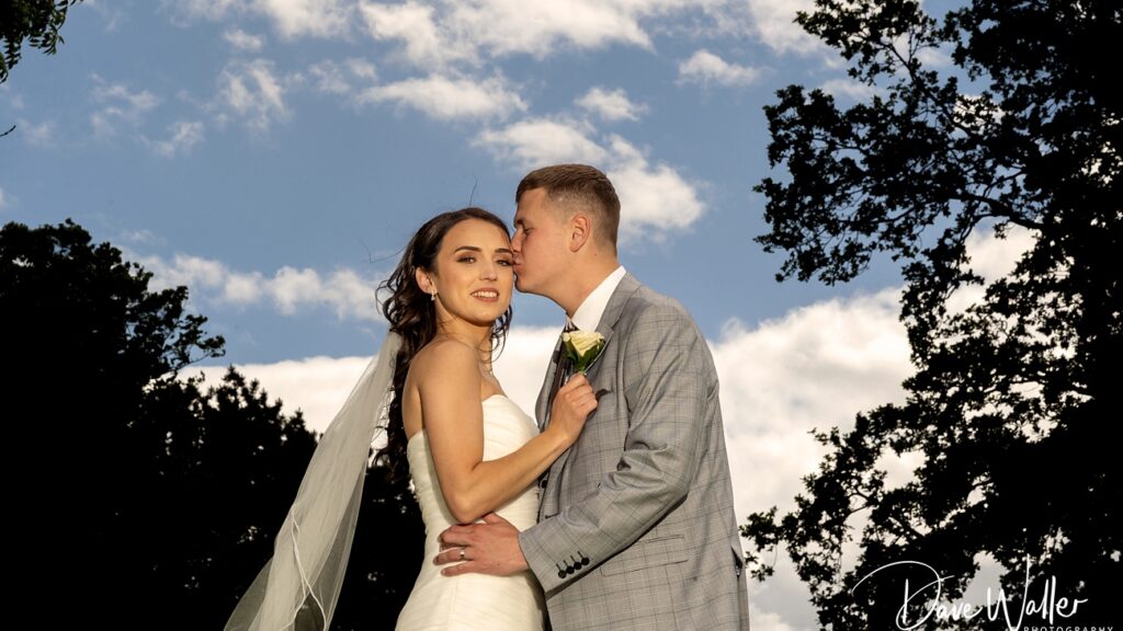 Bride and groom embracing outdoors, sunny sky with trees.