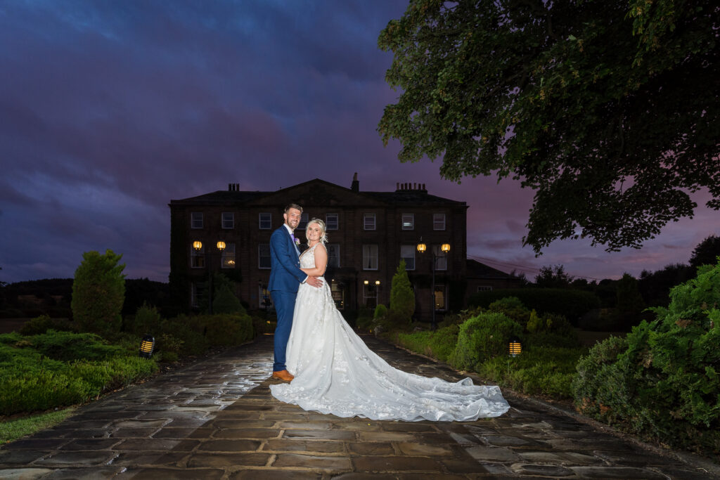 Couple posing at twilight, elegant wedding attire, manor backdrop.