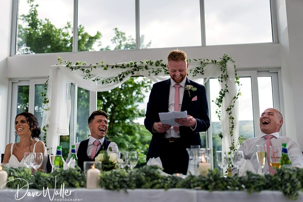 Groom giving speech at wedding, guests laughing.