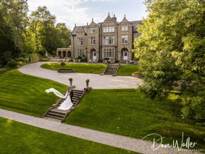 A bride in a flowing white gown descends stone steps in a lush garden with manicured lawns leading to an elegant, large stone house, under a sunny sky in Nottinghamshire.