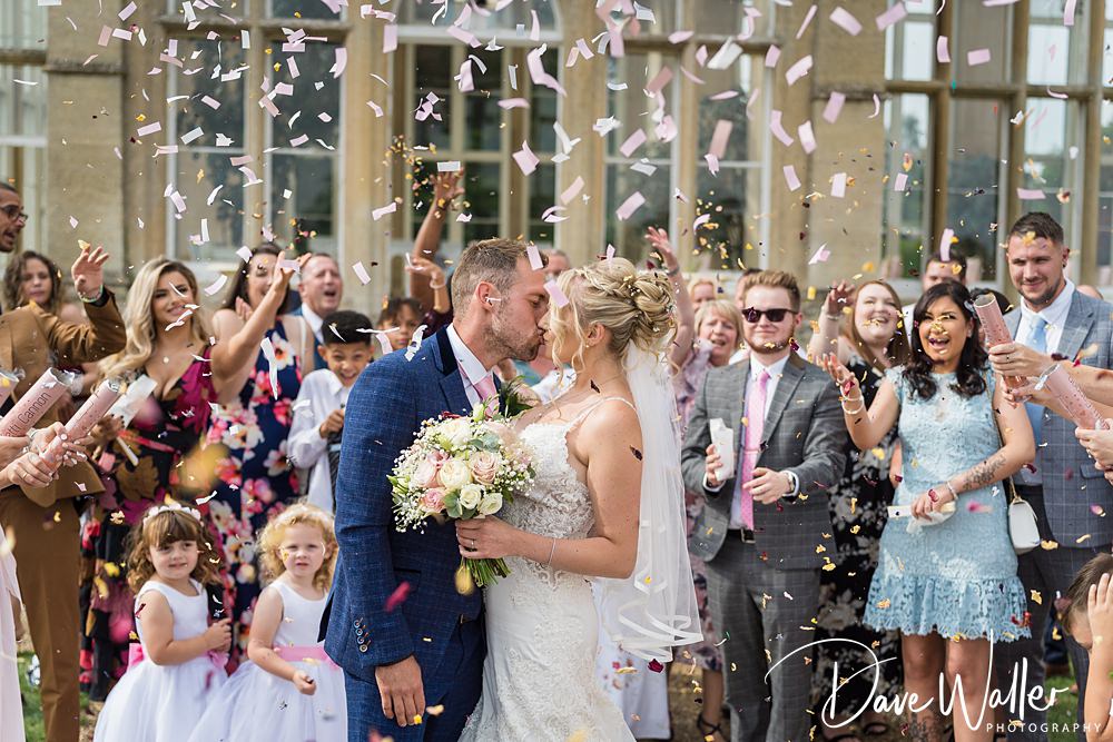 A bride and groom kiss amidst a joyful crowd and a shower of confetti outside a building in Nottingham, with guests cheering and clapping around them.
