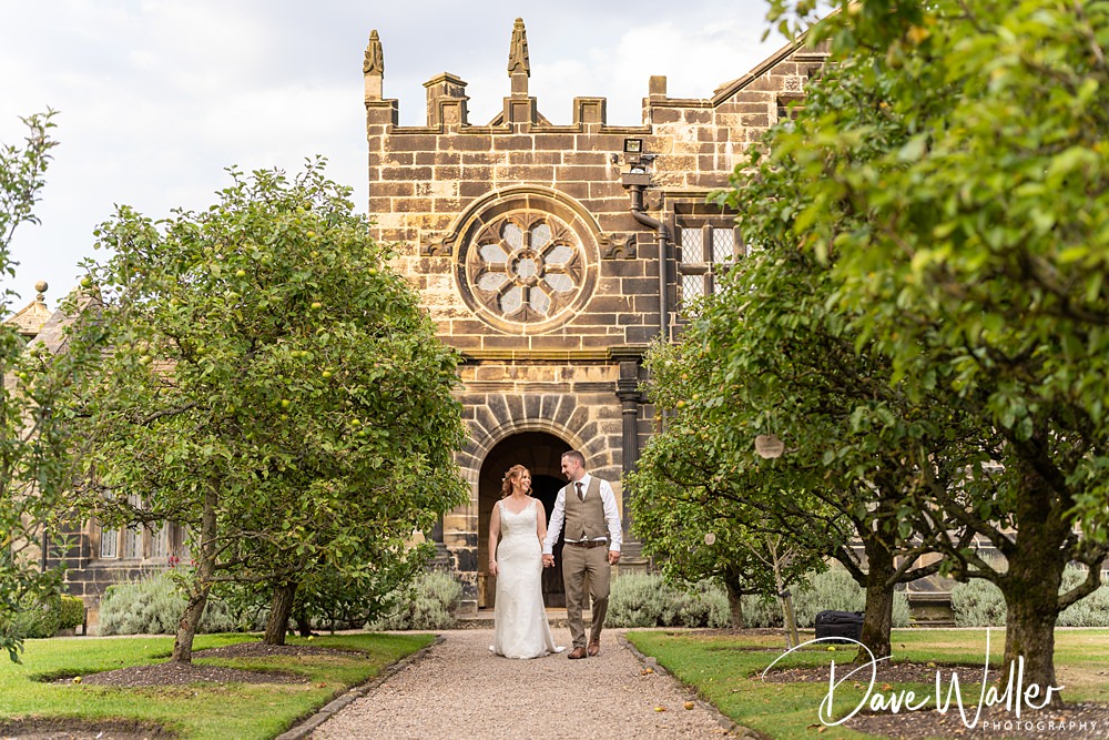 A bride and groom walk hand in hand down a path lined with green trees in Nottingham, with a large, ornate stone building featuring a circular window in the background.