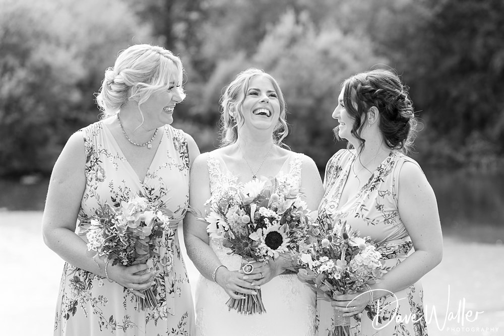 Three women in floral dresses laugh joyously at a Nottingham wedding, each holding a bouquet. They stand outdoors with a blurred natural backdrop, in a black and white photo.