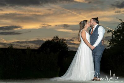 A bride and groom share a kiss at dusk under a dramatic sky, with the bride in a flowing white gown and the groom in a grey suit, highlighted by a soft backlight. Captured by a