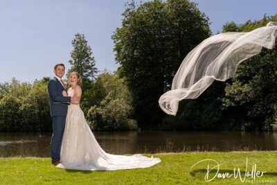 A bride and groom stand by a pond, holding each other and smiling. The bride's veil is tossed by the wind, creating a sweeping arc above them. Lush trees and clear skies in the
