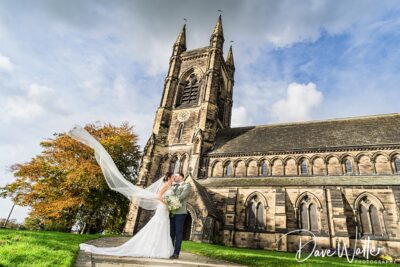 A bride and groom kiss outside a grand stone church with a tall steeple in Nottingham, under a clear blue sky. The bride's long veil flows elegantly in the breeze.