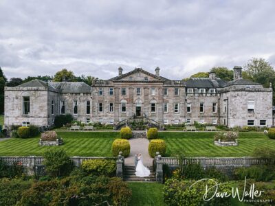 A bride and groom stand at the end of a symmetrical garden pathway leading to an elegant, classic mansion with lush greenery under a cloudy sky, beautifully captured by a York Wedding Photographer.