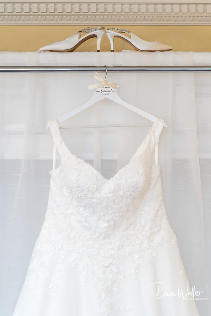 An elegant white wedding dress with lace details hangs from a personalized hanger labeled "JOANNE & BRIAN's Wedding," showcased in front of a softly lit sheer curtain backdrop at Bagden Hall