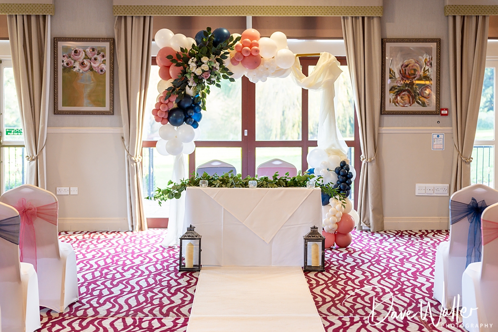 An elegant wedding ceremony setup at Bagden Hall with a white aisle runner, decorated chairs, and a floral arch over the altar, inside a room with large windows and a patterned carpet.