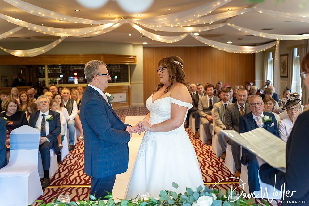 JOANNE & BRIAN's Wedding: A bride and groom hold hands during their wedding ceremony, surrounded by guests in a venue adorned with ceiling fairy lights and floral decorations.