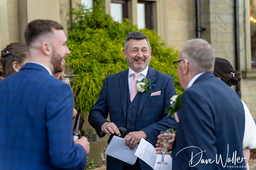 A cheerful man in a blue suit with a boutonniere smiles during a conversation with guests holding drinks at an outdoor wedding event, with the photographer's watermark in the corner.