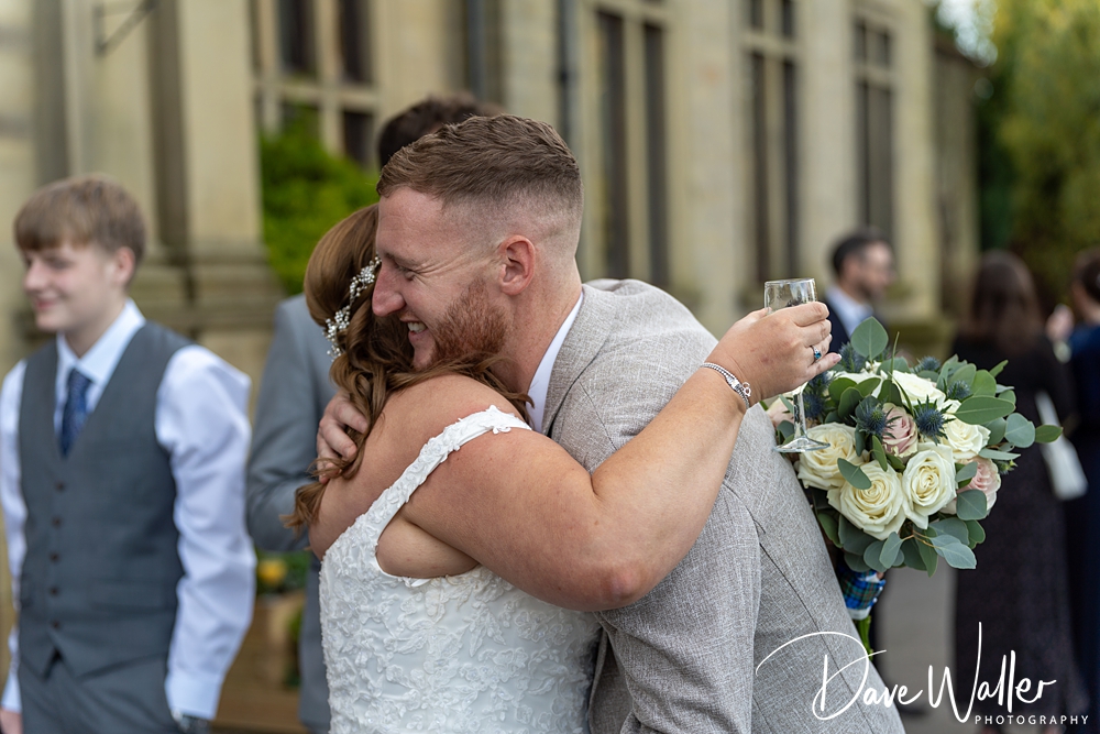 A joyous bride and groom, Joanne and Brian, embracing and laughing outside at their Bagden Hall wedding, with guests in the background. The groom holds a champagne glass and the bride carries a