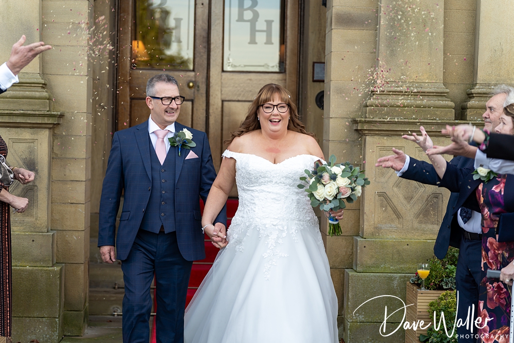 A joyful bride and groom, JOANNE & BRIAN, walking out of Bagden Hall while guests throw confetti over them, both smiling widely. The setting features classic architecture.