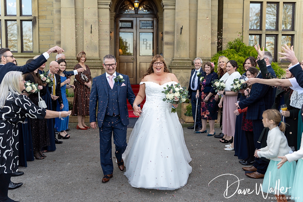 A newlywed couple, Joanne and Brian, joyfully walks through a crowd of guests throwing flower petals, while everyone around them is smiling and clapping, outside the grand historic Bagden Hall.